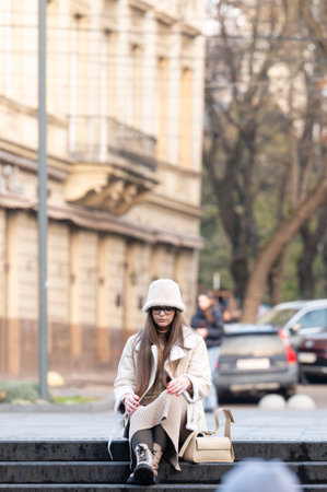Elegant young woman sits peacefully on steps, dressed in a fashionable pale coat and hat, with the lively backdrop of city buildings and people in softly lit afternoon.の写真素材