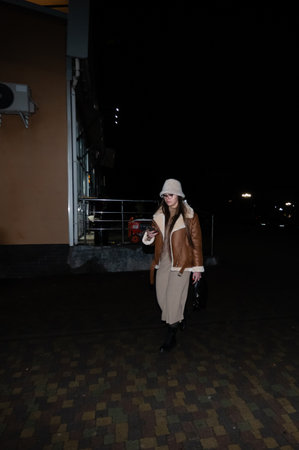 A stylish individual dressed in a warm brown jacket and light dress strolls confidently down a street under the glow of nearby lights, exuding elegance and graceの写真素材