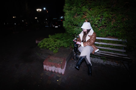 Under the glow of streetlights, a fashionable individual sits on a park bench, dressed in winter attire, enjoying a quiet moment surrounded by lush greenery.の写真素材