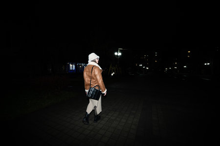 Under the glow of streetlights, a person in a brown leather jacket and white hoodie strolls along a cobblestone path, embracing the tranquility of a nighttime city atmosphere.の写真素材