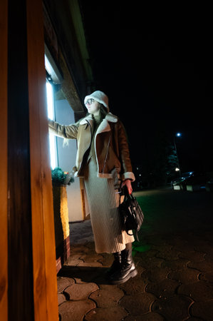 Under a starry night, a fashionable individual interacts with a softly lit storefront, showcasing their winter style amidst a quiet urban environment.の写真素材