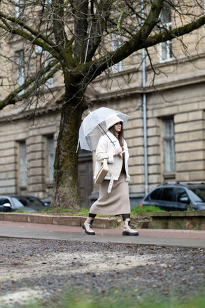A woman dressed in a stylish outfit strolls leisurely along a street adorned with classic buildings while holding a clear umbrella, embracing the rainy weather.の写真素材