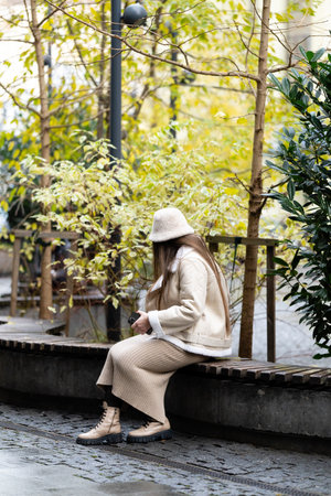 A woman dressed in stylish beige clothing relaxes on a bench surrounded by lush foliage in a calm urban park, embracing the serene atmosphere of the afternoon.の写真素材