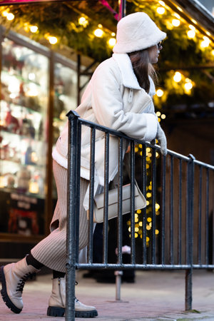 A woman dressed in a cozy winter coat and hat leans against a railing, surrounded by sparkling holiday lights at a lively city spot, creating a serene seasonal vibe.の写真素材