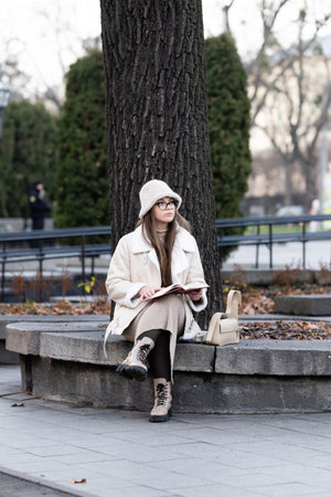 Wrapped in warm layers, a young woman sits on a stone bench beneath a large tree, immersed in her book amidst the tranquil atmosphere of a late autumn afternoon.の写真素材