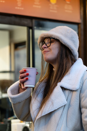 A woman with long brown hair wearing a cozy white coat and a fluffy hat enjoys a warm drink, smiling as she savors the moment in a bustling urban setting.の写真素材
