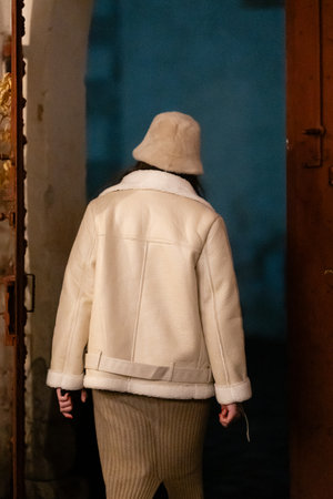 A stylish individual in a warm white coat and beige hat stands at a wooden door, capturing a moment of fashionable solitude in a softly lit environment.の写真素材