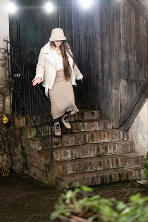 Charming courtyard ambiance sets the stage as a woman, warmly dressed, navigates brick steps with elegance under twinkling lights during the cool evening.の写真素材