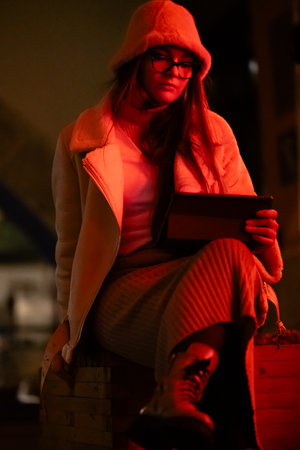 In a vibrant city atmosphere, a young woman dressed in a soft coat and warm boots sits comfortably, immersed in her tablet while night lights glow around her.の写真素材