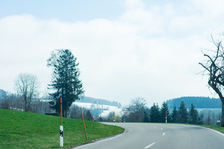 Beneath a canopy of soft clouds, a gentle curve in the road leads through vibrant grass and picturesque hills, heralding the arrival of spring in the countryside.の写真素材