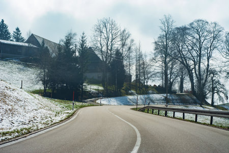 A serene winter day reveals a winding road bordered by white snow and tall trees, leading to charming homes nestled along the hillside, creating a picturesque, peaceful atmosphere.の写真素材