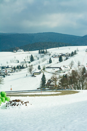 A vibrant green sculpture stands in a snowy field, overlooking a picturesque village nestled among rolling hills, creating a striking contrast against the winter scenery.の写真素材