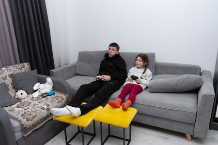 Two siblings sit comfortably on a stylish couch, engrossed in an exciting gaming session. Their...
