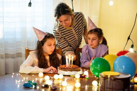 Two girls wearing festive hats smile as they prepare to blow out birthday candles on a cake,...