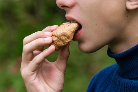 Close-up of a person wearing a blue turtleneck sweater taking a bite of a small, golden-brown pastry. The blurred green background suggests a natural outdoor setting.の写真素材