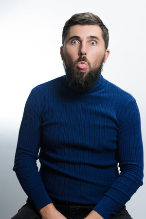 A bearded man in a blue turtleneck sticks his tongue out against a white background. His playful expression conveys humor and lightheartedness.の写真素材