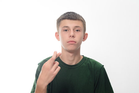 A young man with short brown hair, wearing a dark green t-shirt, sits against a white background. He's making a hand gesture, fingers crossed, suggesting a wish or hope.の写真素材