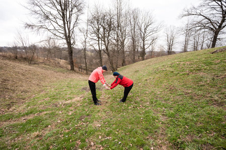 Friends engage in a joyful, shared stretching exercise amidst a tranquil natural setting, surrounded by rolling hills and bare trees on a cool spring day.の写真素材