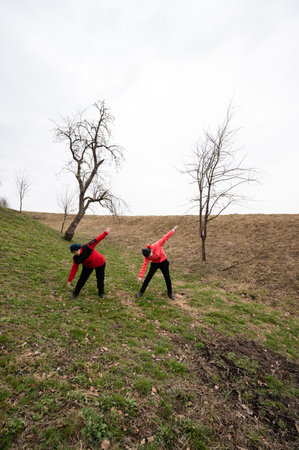 Two individuals dressed in vibrant red stretch their arms in unison on a grassy slope beneath a hazy sky, surrounded by sparse, leafless trees.の写真素材