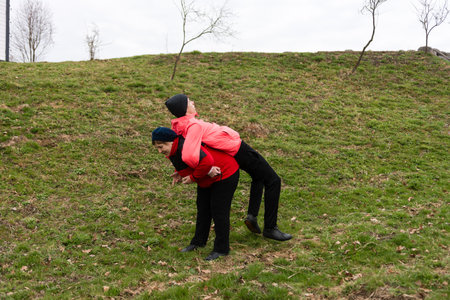 Friends share a joyful moment, with one giving a piggyback ride while surrounded by lush greenery in a peaceful outdoor environment under overcast skies.の写真素材