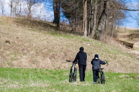 Two individuals, possibly father and son, stroll along a grassy path. Bicycles in tow. Serene countryside setting, bathed in sunlight. Hills and trees surround them.の写真素材