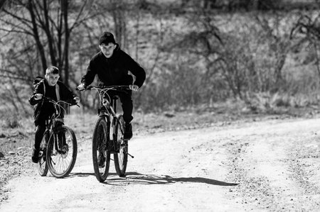 Two young boys are cycling down a country lane. The black and white photo shows them enjoying a ride through nature on a sunny day. The path is dirt, and trees line the background.の写真素材