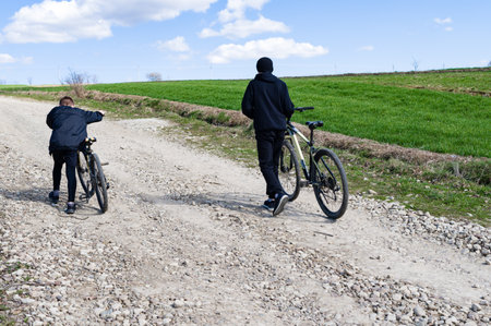 Two young people walk their bicycles along a gravel path. The path winds through a countryside scene of green fields under a partly cloudy sky.の写真素材
