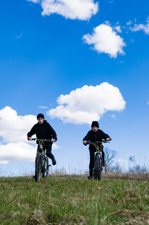 Two young people on bicycles ride uphill on a sunny day. The sky is a brilliant blue with puffy white clouds. Grass covers the hill.の写真素材