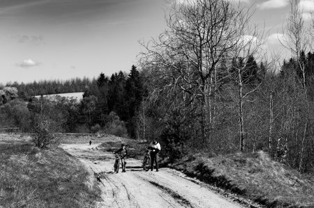 A black and white photo of two people on bicycles taking a break on a country road. Trees line the path, creating a serene, natural setting. The atmosphere is peaceful and quiet.の写真素材