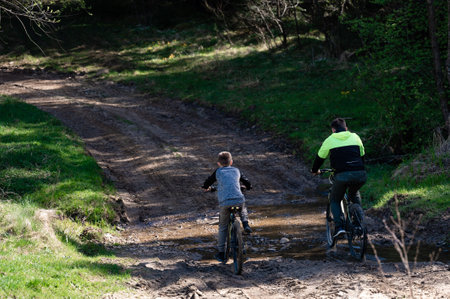 Two figures on bicycles traverse a muddy path in a woodland setting. The older individual leads the younger, creating a heartwarming scene of shared outdoor activity.の写真素材