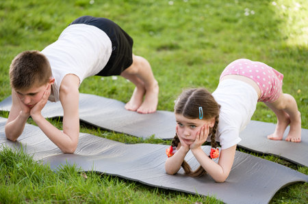 Two children, a boy and a girl, perform yoga poses on grey mats in a grassy area. They are resting...