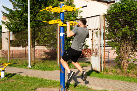 A man performs pull-ups on outdoor exercise equipment. He's wearing athletic attire in a park-like...