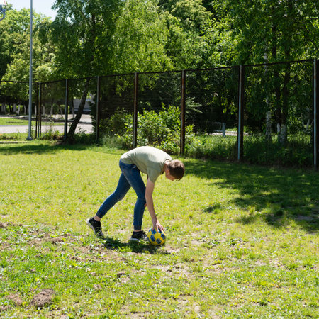 A young person in casual clothes retrieves a soccer ball from a grassy outdoor area near a fence. Sunlight illuminates the scene.の写真素材