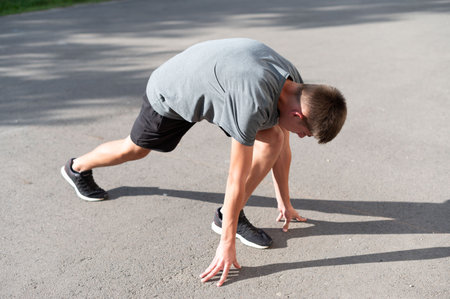 A young man in athletic wear performs a runner's stretch on asphalt. Sunlight casts shadows on the...