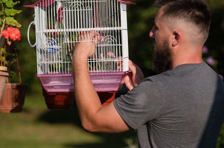 A bearded man carefully holds a birdcage containing a small bird. The scene is set outdoors in a garden-like setting, bathed in sunlight.の写真素材