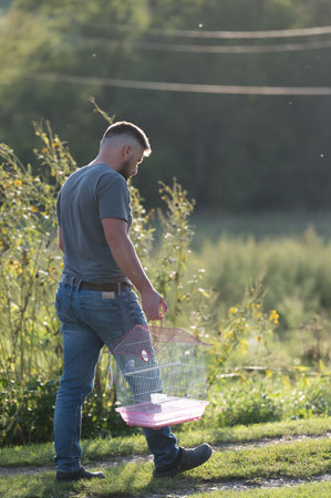 A man walks along a path, carrying a small pink birdcage. The setting is a rural area with green grass and wildflowers. The sun shines brightly, casting a warm light.の写真素材