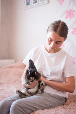 A heartwarming scene of a young woman lovingly cradling a dark-furred rabbit. She sits on a plush bed, her gentle touch conveying care and affection for the pet.の写真素材