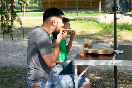 A man and boy sit at a small folding table in a park, eating pizza slices and drinking juice. The scene is relaxed and joyful, showcasing a heartwarming father-son moment.の写真素材
