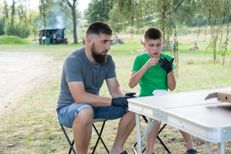 A man and boy sit at a portable table in a park, preparing food while wearing black gloves. They appear relaxed and enjoying a picnic-style meal.の写真素材