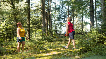 An adult woman and a young child enjoy a game of catch in a lush forest setting. Sunlight filters...