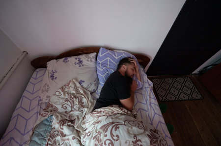 A man with a beard is asleep in a bed with patterned sheets and pillows. His head rests on his hand....