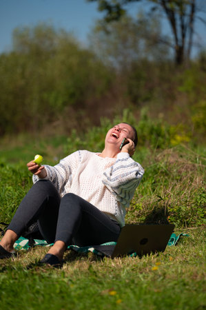 A woman sits in a sunlit meadow, laughing heartily while talking on her phone and holding a half-eaten apple. A laptop rests beside her on a blanket. Trees and bushes form the backdrop.の写真素材