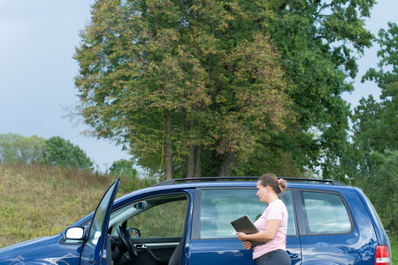 A woman in a pink shirt and gray pants stands in a grassy field next to a blue car with an open door, holding a tablet. Large green trees form a backdrop with a cloudy sky.の写真素材