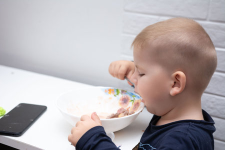 A toddler is seated at a white table, intently eating from a floral patterned bowl using a spoon. The room is well lit, with a white background.の写真素材