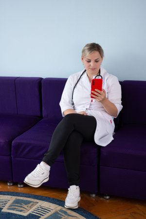 A female doctor wearing a white coat and stethoscope is sitting on a purple couch, looking at her red smartphone. She is wearing black leggings and white sneakers.の写真素材