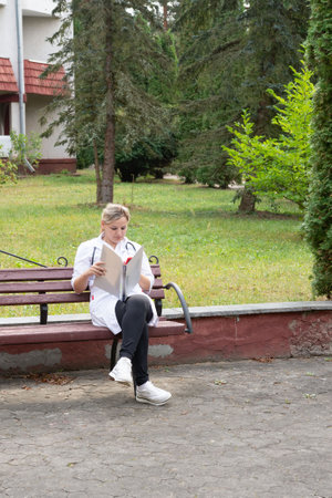 A medical professional in a lab coat is sitting outside on a bench, engrossed in reading materials. The surrounding park offers a tranquil scene for relaxation and review. The ground is paved.の写真素材