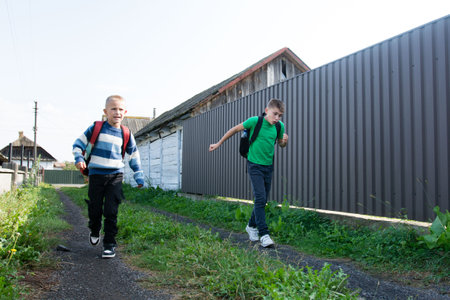In a rural setting, two boys with backpacks are seen running down a grassy, gravel path. They are near fences and buildings with a sense of urgency in their stride. The sky is bright.の写真素材