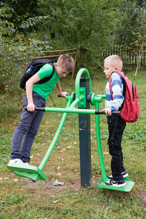 Two young boys are exercising on outdoor fitness equipment in a park. They both are wearing backpacks, suggesting they are students. The scene promotes active lifestyles and outdoor fun.の写真素材