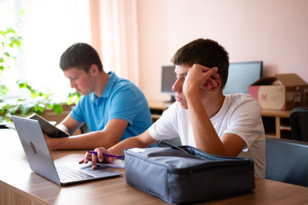 Two young male students are engaged in studying. One is working on a laptop while the other reads....