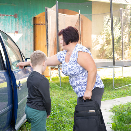 A mature woman stands near a blue car, seeing off a young boy. She holds a black bag. They're in a grassy yard outside an older home with a trampoline visible in the background.の写真素材